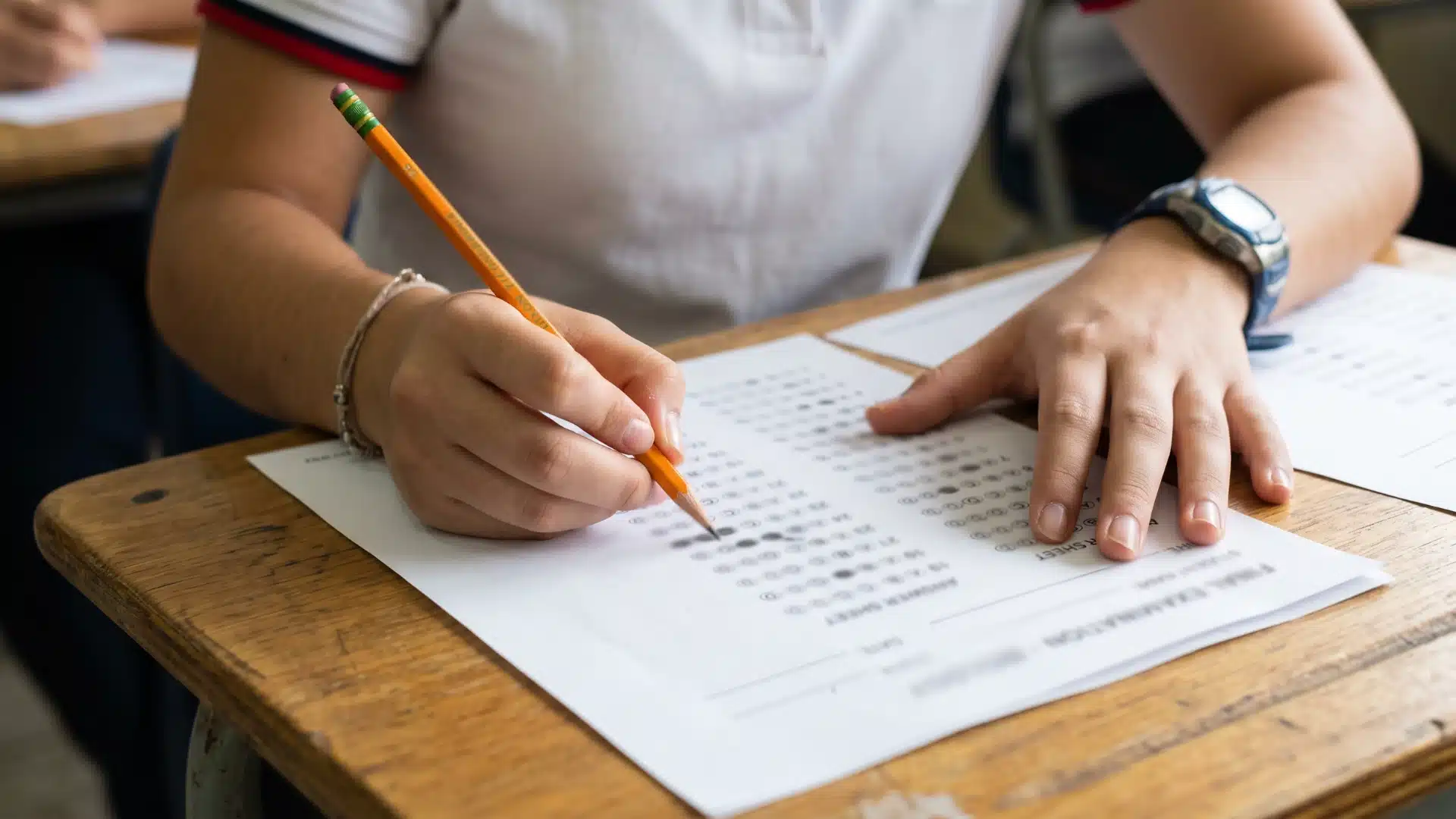student's hands filling out a multiple-choice answer sheet with a yellow pencil while wearing a watch at a wooden school desk