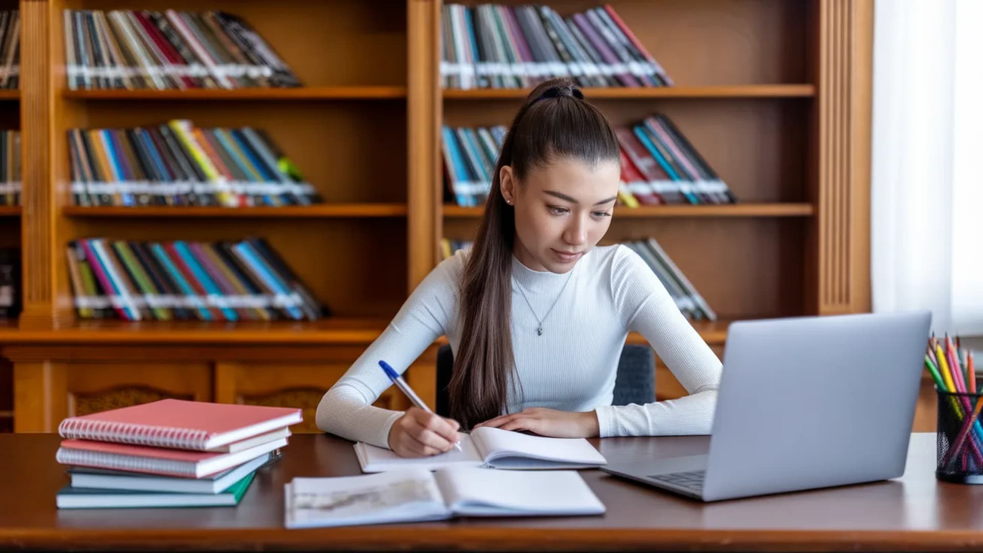 A teenage girl writing notes in a notebook while studying on a laptop in a library.