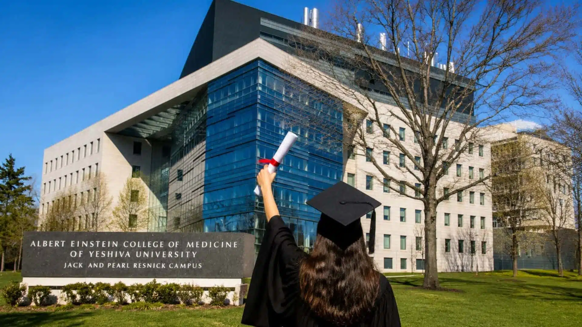graduate in black gown raising diploma on green lawn with modern college building in background under blue sky
