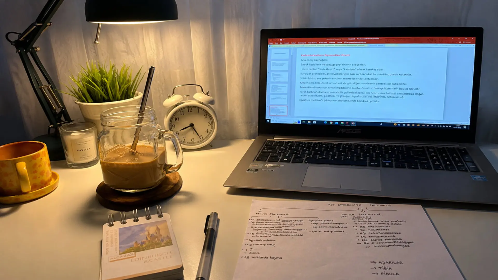 student study desk with laptop, handwritten notes, coffee, lamp, clock, and study materials