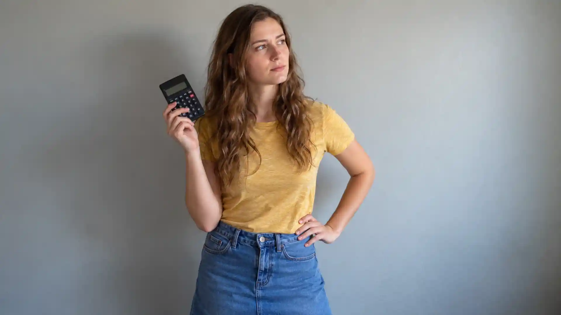 woman in a yellow shirt holding a calculator with a thoughtful expression against a plain wall