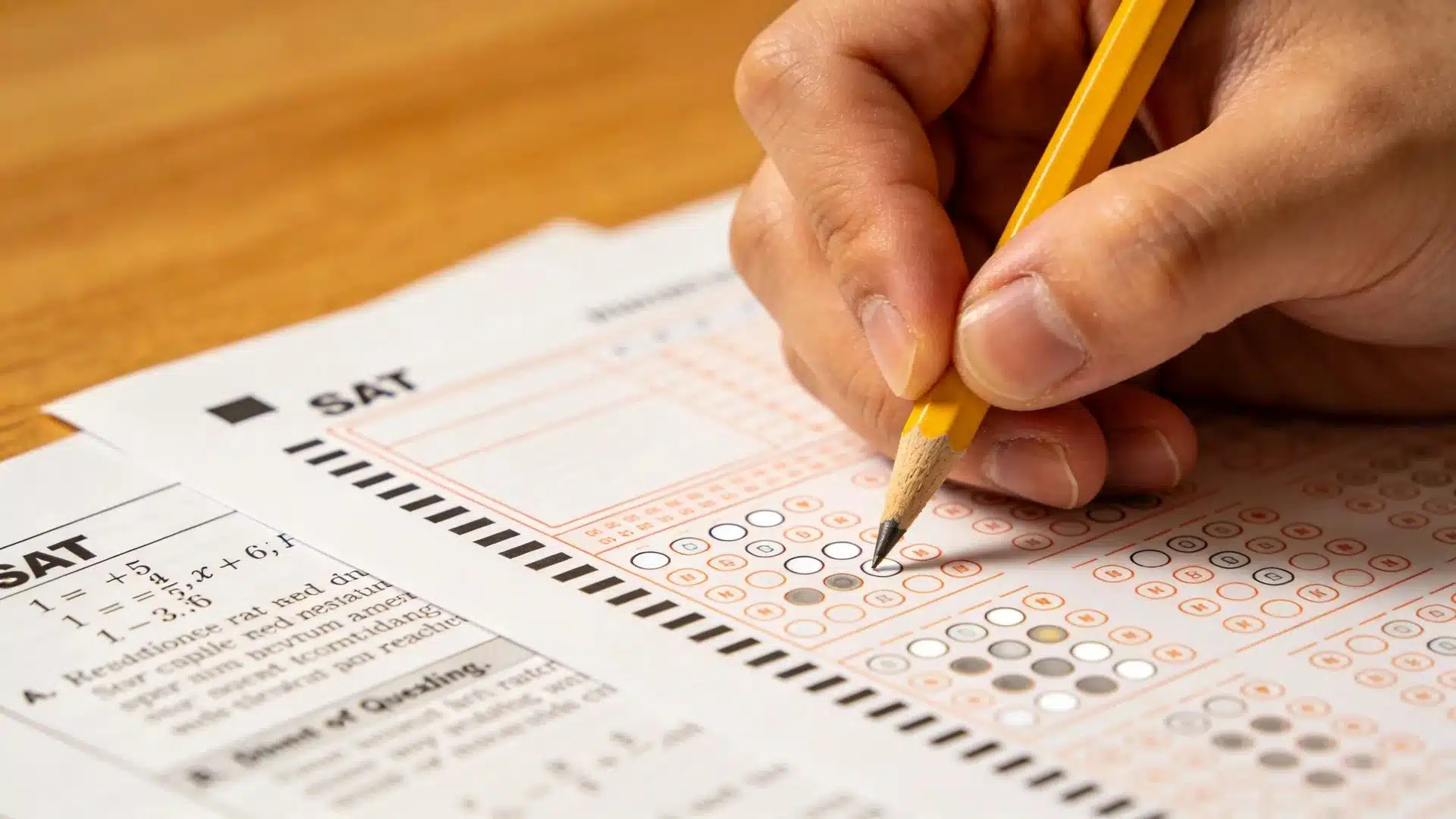 a hand holding a yellow pencil filling in bubbles on an SAT answer sheet on a wooden desk surface