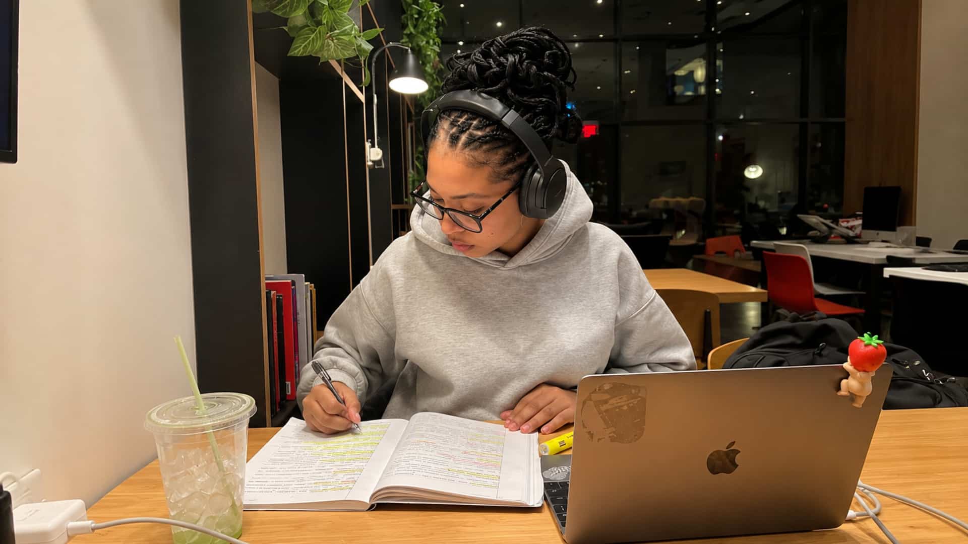 student in headphones highlighting notes in a textbook beside a laptop at night