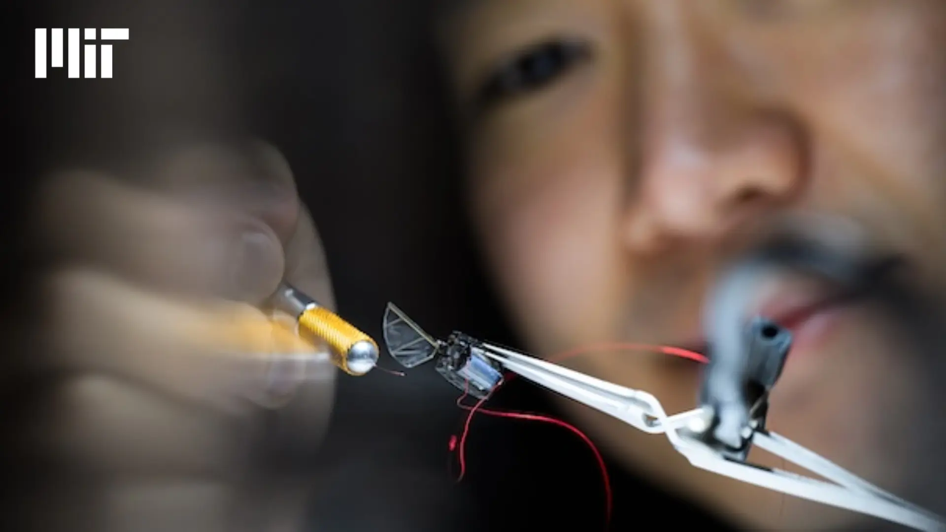 close-up of a soldering iron near a tiny robotic device held by tweezers, blurred face behind, mit logo top left
