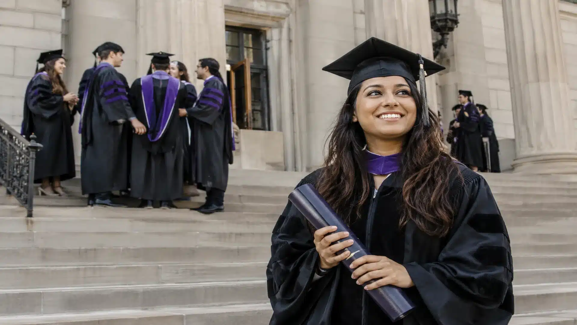 law school graduate holding a diploma outside a courthouse-style campus building with fellow graduates nearby