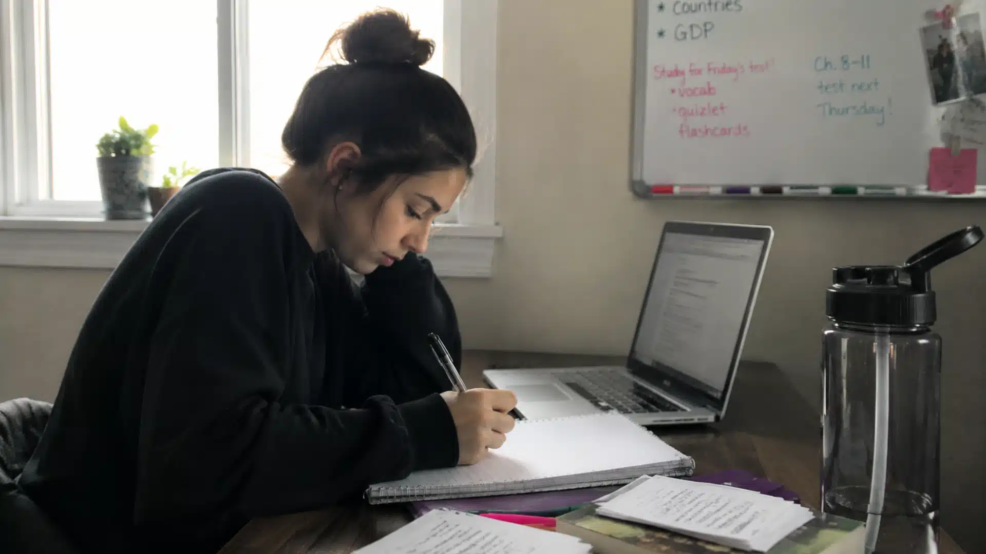 student taking notes beside a laptop while studying for the sat at a quiet desk near a window