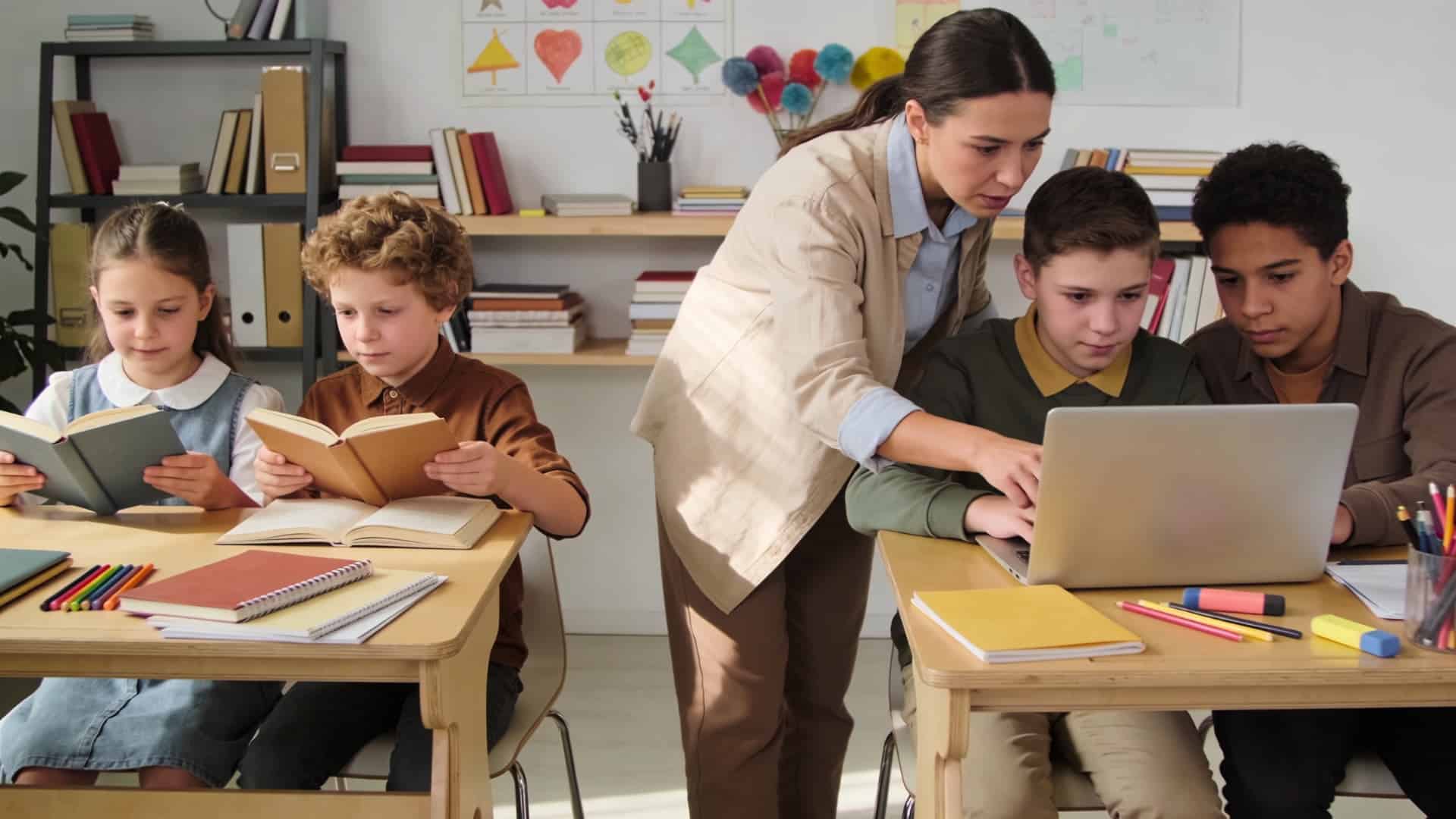 teacher helping students with books and laptop during an interactive classroom lesson with diverse grade levels and subjects